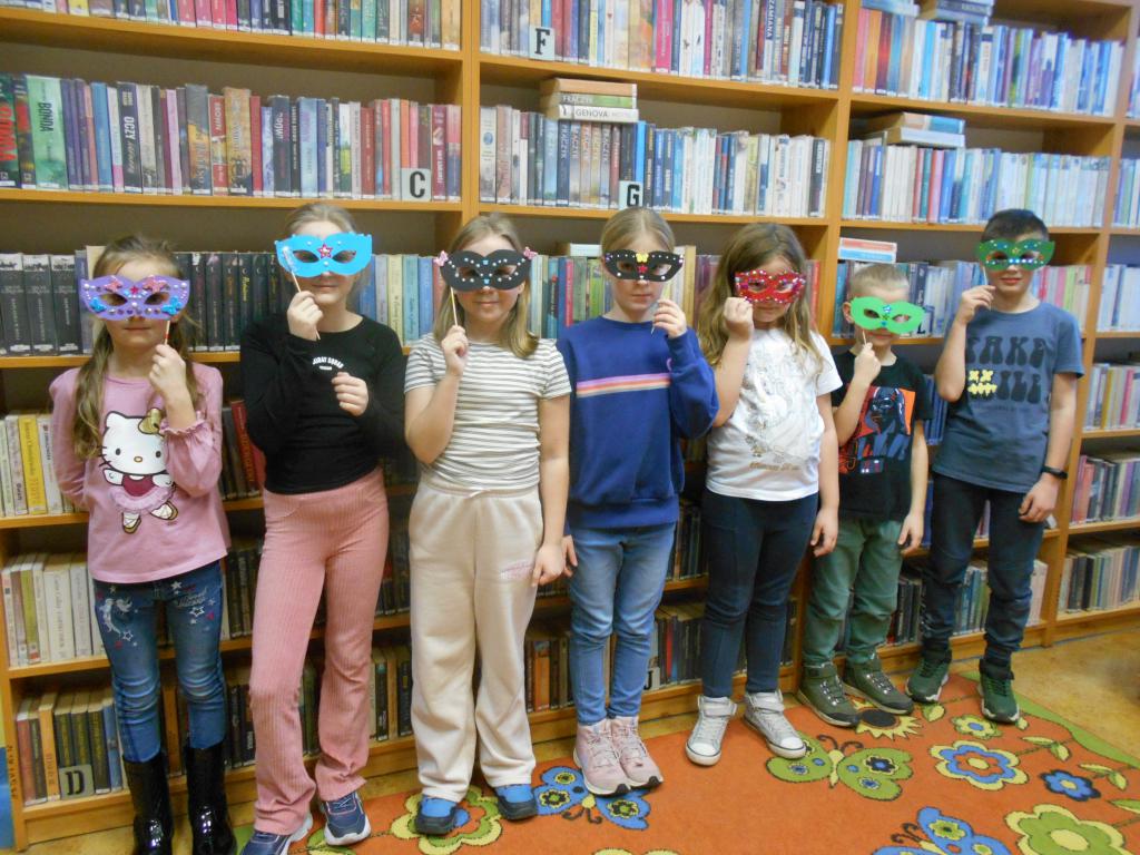 Uczestnicy zajęć stoją pod regałami bibliotecznymi, twarze mają przysłonięte wykonanymi przez siebie maskami karnawałowymi