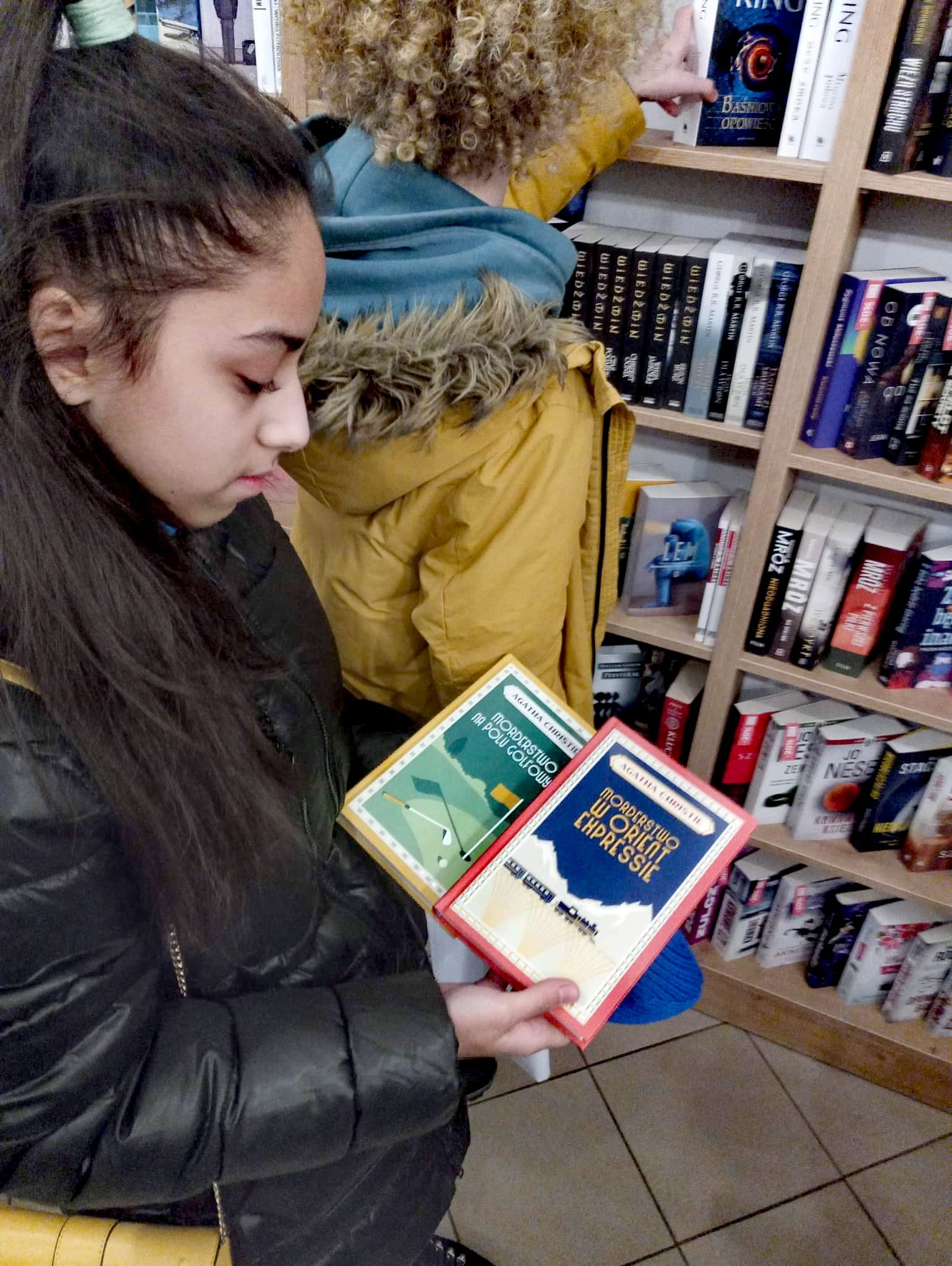 Ciemnowłosa dziewczyna stoi w księgarni i trzyma w rękach dwie książki