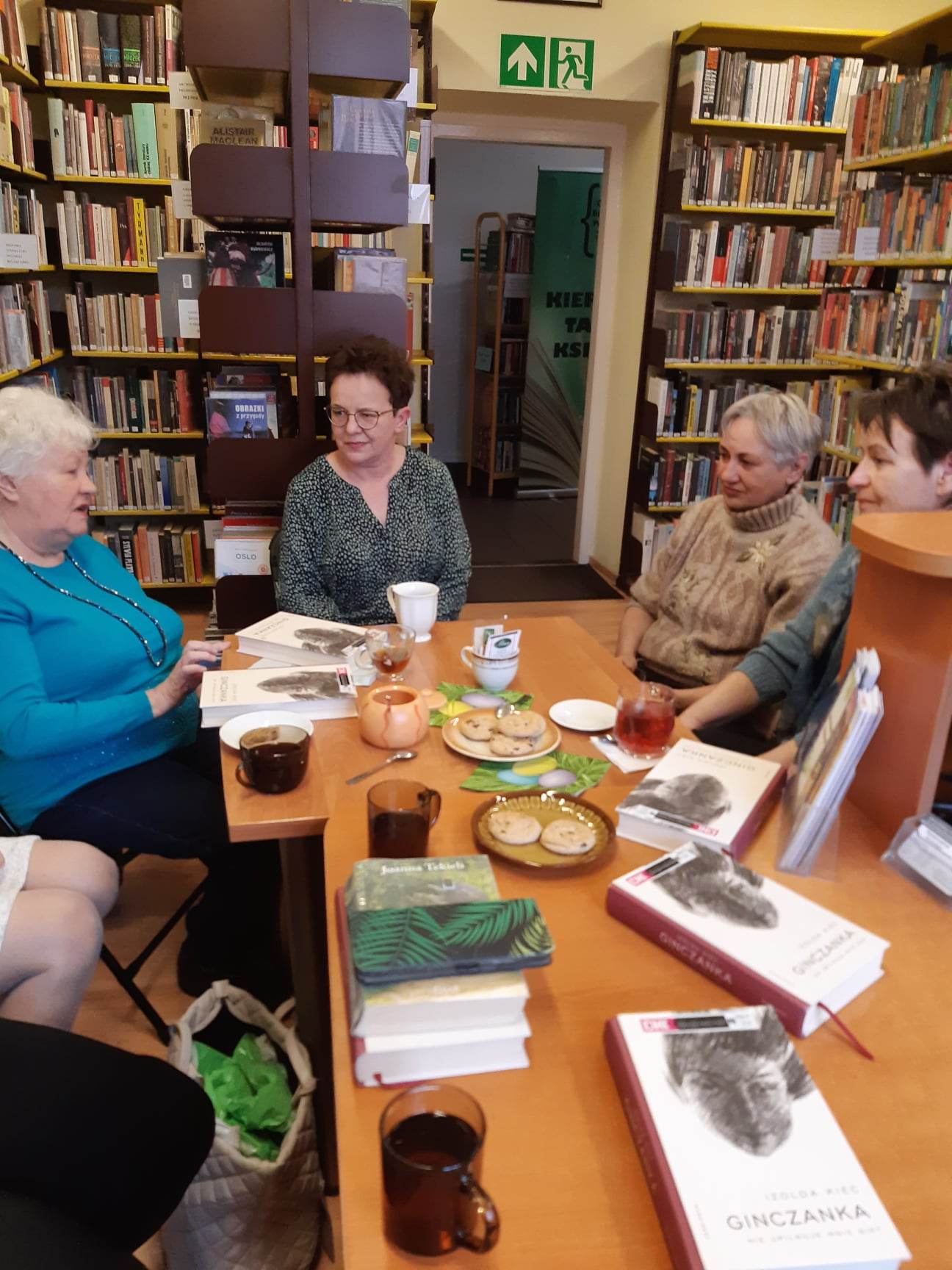 Seniorki siedzą przy stolikach w otoczeniu książek i rozmawiają