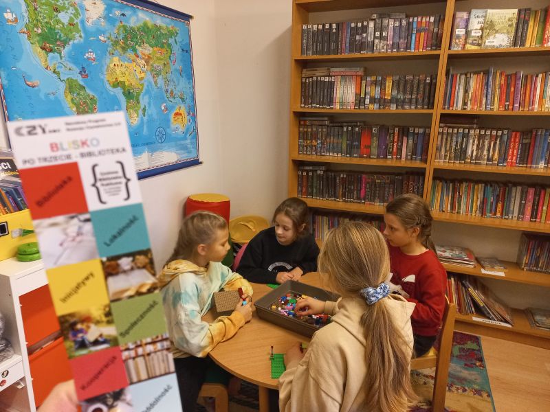 Cztery dziewczynki z wielkim entuzjazmem budują z klocków lego przy bibliotecznym stoliku